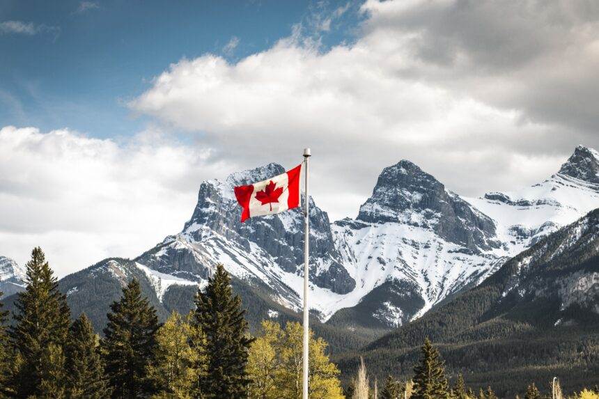 A nature view with big mountains and trees and a tall Canadian flag flaring in the middle. The concept of what is Canada known for.