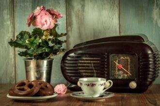 A vintage radio on a table with a flower vase, cookies and a tea cup.